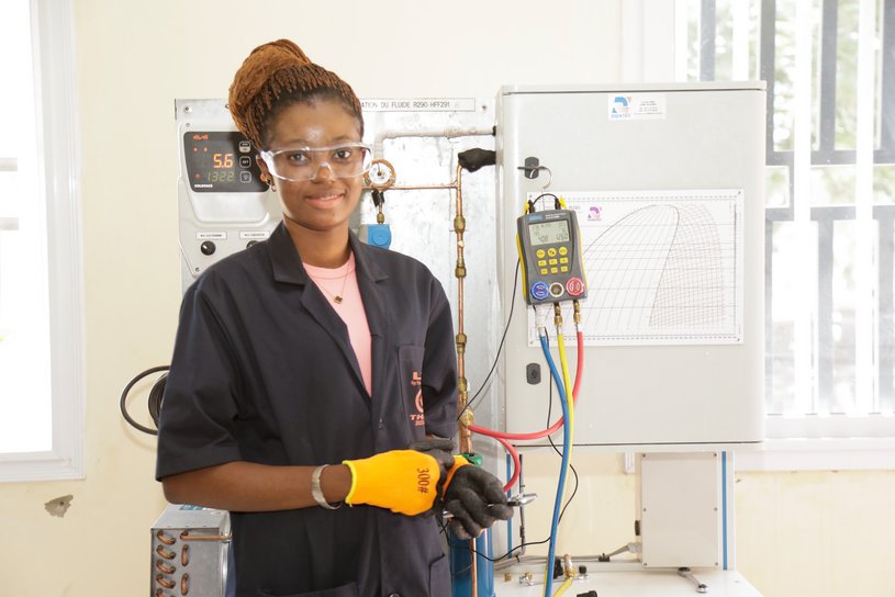 [Translate to French:] A young women with protective clothing stands in front of a training bench for natural refrigerants. She smiles.